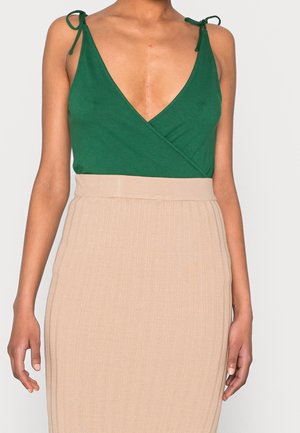 Femme portant un haut cache-cœur vert sans manches avec des bretelles nouées aux épaules et une jupe beige côtelée taille haute, torse visible sur fond uni.