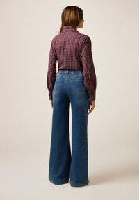 Bordeaux geruit shirt met lange mouwen, gecombineerd met wijde blauwe jeans met metallic versieringen langs de zijkanten.
