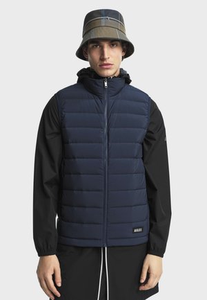 Jeune homme portant une veste matelassée bleue sur une veste à capuche noire et un chapeau seau à carreaux verts, face à l'avant avec une expression neutre.