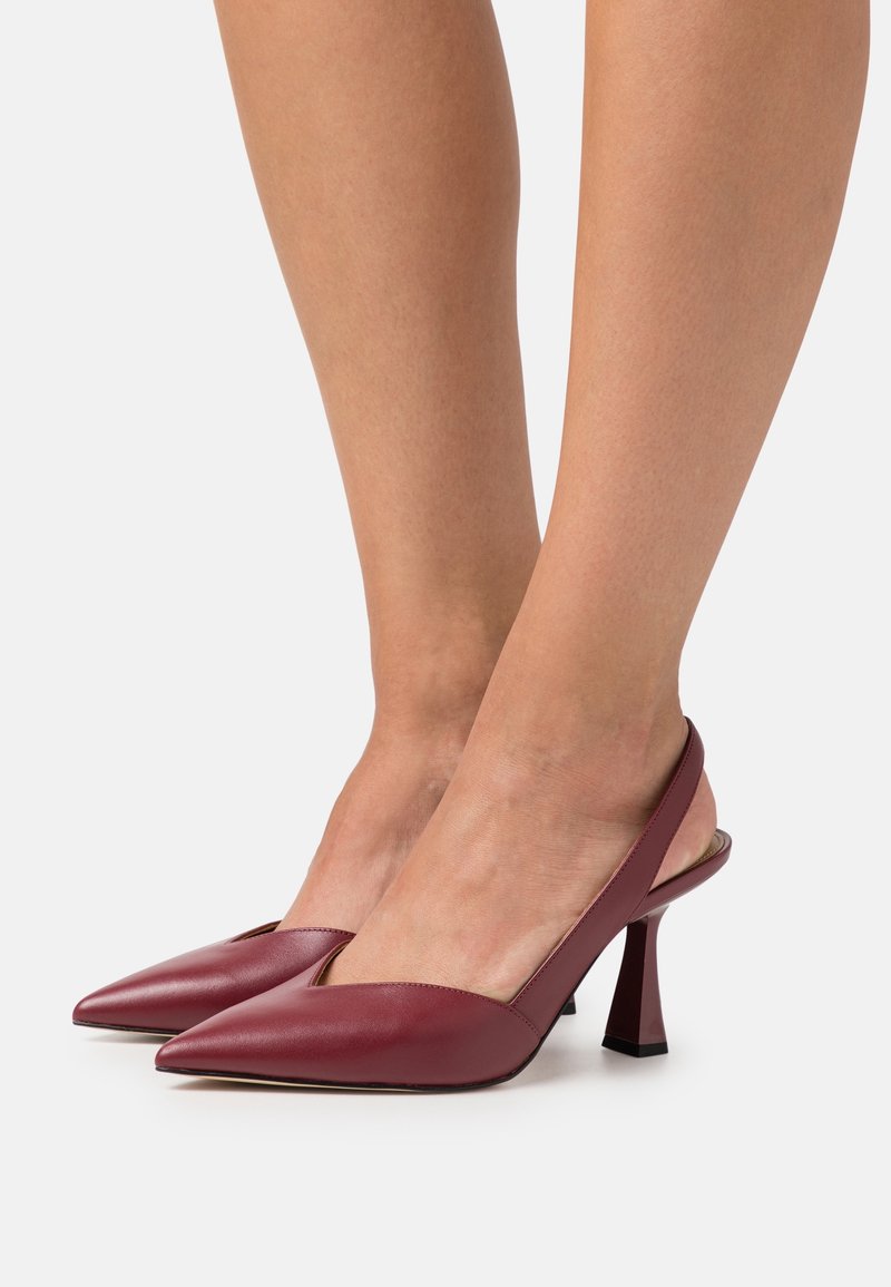 Jambes de femme portant des escarpins slingbacks bordeaux à bout pointu avec talons carrés, sur un fond blanc uni.