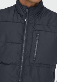 Gilet matelassé bleu marine avec col montant, doté d'une fermeture éclair à l'avant et d'une poche poitrine zippée. Tissu lisse avec un aspect matelassé.