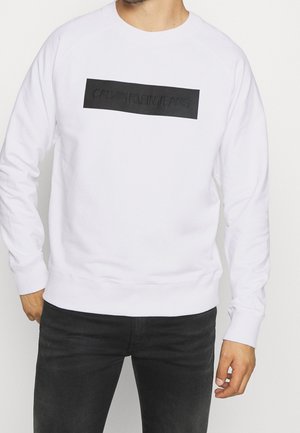 Man draagt een wit sweatshirt met lange mouwen met een zwarte "CALVIN KLEIN JEANS" logo in blokletters op de borst, gecombineerd met een zwarte broek.