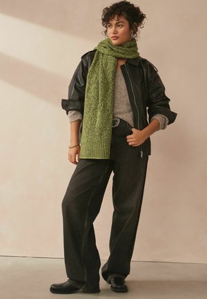 Grøn strikket tørklæde lagd over en grå sweater, parret med en sort læderjakke og brede mørkeblå jeans. Sorte sko fuldender looket.
