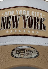 Μπεζ και λευκό καπέλο με τα κέντηματα "NEW YORK CITY" και "NEW YORK" σε καφέ χρώμα. Διαθέτει πολλούς λευκούς αστέρες, ρυθμιζόμενο λουράκι και ετικέτα με το σήμα.