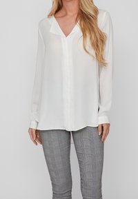 Blusa bianca a maniche lunghe con scollatura a V, realizzata in tessuto trasparente, dotata di chiusura con bottoni e polsini. Abbinata a pantaloni slim-fit grigi a quadri.