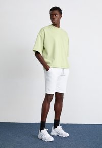 Světle zelené oversized tričko, bílé šortky a bílé sportovní boty s síťovinovým vzorem. Model má krátké tmavé vlasy a stojí na modrém koberci.