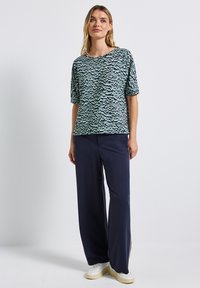 Nainen seisoo päällään vihreä-musta kuviollinen lyhythihainen paita, tummansiniset leveälahkeiset housut ja valkoiset lenkkarit yksinkertaista taustaa vasten.