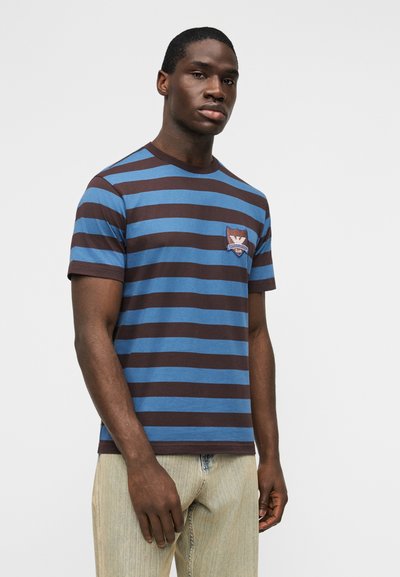 Άνδρας που στέκεται φορώντας μπλε και καφέ οριζόντια ριγέ μπλούζα με μικρό λογότυπο στο στήθος και ανοιχτόχρωμο τζιν, ουδέτερο φόντο.