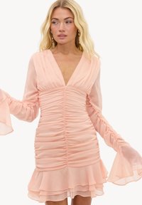 Femme blonde en robe longue rose pâle à manches longues en tissu transparent avec un ourlet à volants superposés et des poignets évasés, regardant légèrement sur le côté.