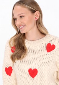 Cremfarbener Strickpullover mit mehreren roten Filzherz-Applikationen. Besitzt einen gerippten Rundhalsausschnitt und einen lockeren Schnitt. Glattes Haar, das über die Schultern fällt.