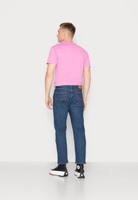 T-shirt rosa de manga curta, jeans de denim azul com bolsos traseiros e um patch de pele com marca, sapatilhas altas pretas com solas brancas.