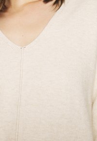 Maglione beige a V con un tessuto morbido e testurizzato, caratterizzato da un sottile dettaglio a cucitura lungo il centro anteriore.