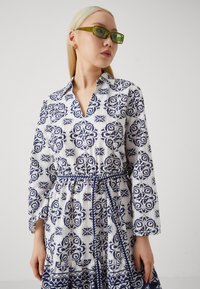Rochie albă cu un model floral albastru, guler la gât, maneci lungi și talie strânsă cu o panglică asortată. Textură de material neted.