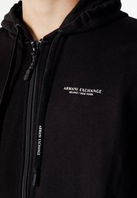 Haut à capuche zippé noir en tissu doux, présentant un logo sur la poitrine et des cordons de serrage. Surface texturée avec des détails de fermeture éclair contrastants.