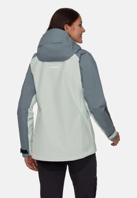 Lys grå udendørsjakke med en mørkere grå hætte og ærmer. Har justerbare manchetter og et subtilt logo på ryggen. Vandtæt stof.