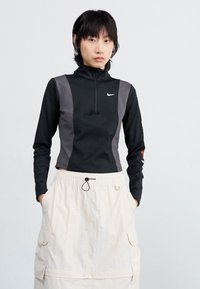Top sportiv negru cu fermoar la jumătate, având panouri gri și logo-ul Nike, asortat cu o fustă crem cu șnur, ce dispune de buzunare și un material texturat.