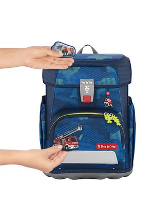 Rucksack im blauen Camouflage-Muster, mit einer Fronttasche im Design eines Feuerwehrautos, grünen Akzenten und einem grauen Boden. Zwei Hände halten zusätzliche Applikationen.
