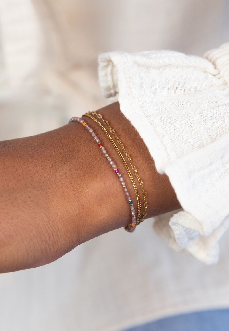 Tre braccialetti su un polso: una catena d'oro, uno con piccoli bead colorati e un delicato filo con sottili accenti iridescenti.