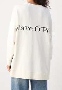 Kremowy, oversize'owy sweatshirt z czarnym nadrukiem "Marc O'Polo" na plecach, z prążkowanymi mankietami i miękką teksturą.