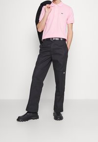Camisa de polo rosa con cuello y un logo verde, acompañada de pantalones negros. Botas negras con suelas texturizadas, blazer informal colgado de un hombro.