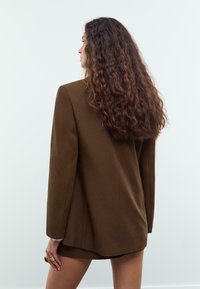 Blazer marron surdimensionné avec une texture lisse, des revers crantés et une coupe droite. Le mannequin est tourné, montrant la vue de dos.