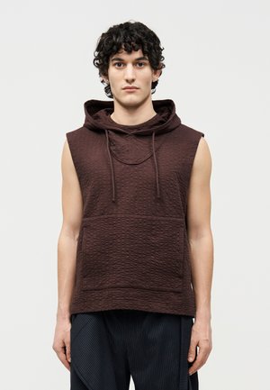 Νέος άντρας με σγουρά μαλλιά, που φοράει ένα αμάνικο, υφασμένο σκουρόκαφέ φούτερ με κουκούλα και σκούρο παντελόνι με ρίγες, στέκεται μπροστά σε απλό φόντο.