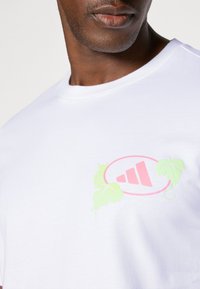 Hvid bomuld t-shirt med et pink logo og grønne vinranker på brystet. Korte ærmer, rund halsudskæring.