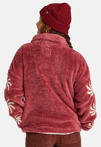 Giacca in pile rosa soffice con colletto alto, caratterizzata da motivi a spirale color crema sulle maniche. Abbinata a un berretto di maglia bordeaux.