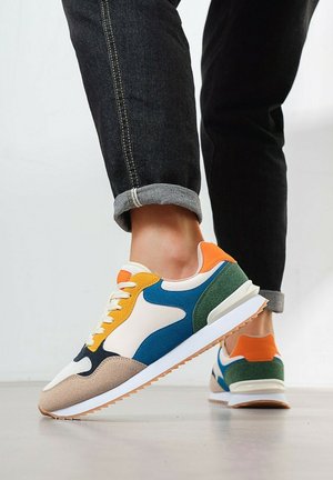 Gros plan sur une personne portant des baskets multicolores avec des accents beige, bleu, vert, orange et un jean noir retroussé, marchant sur un sol clair.