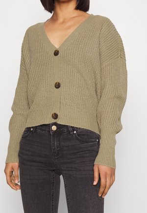 Femme portant un cardigan court en maille vert olive avec trois boutons, associé à un jean skinny noir délavé taille haute.