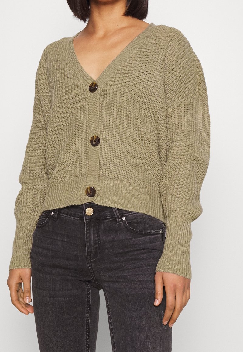 Femme portant un cardigan court en maille vert olive avec trois boutons, associé à un jean skinny noir délavé taille haute.