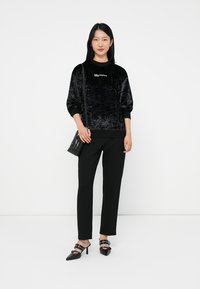 Donna che indossa un maglione di velluto nero, pantaloni neri su misura, mules con tacco neri e porta una borsa a tracolla nera, in piedi su uno sfondo bianco.