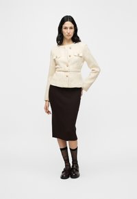 Sötét hajú nő krémszínű, texturált, gombos kabáttal és övvel, fekete midi szoknyában, áttetsző fekete zokniban és sötét loaferekben, magabiztosan állva.