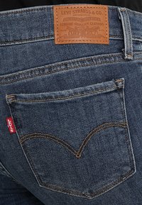 Los jeans de denim azul de Levi's presentan un parche de cuero en la parte trasera, costuras amarillas y una etiqueta roja en el bolsillo trasero, mostrando un diseño clásico.
