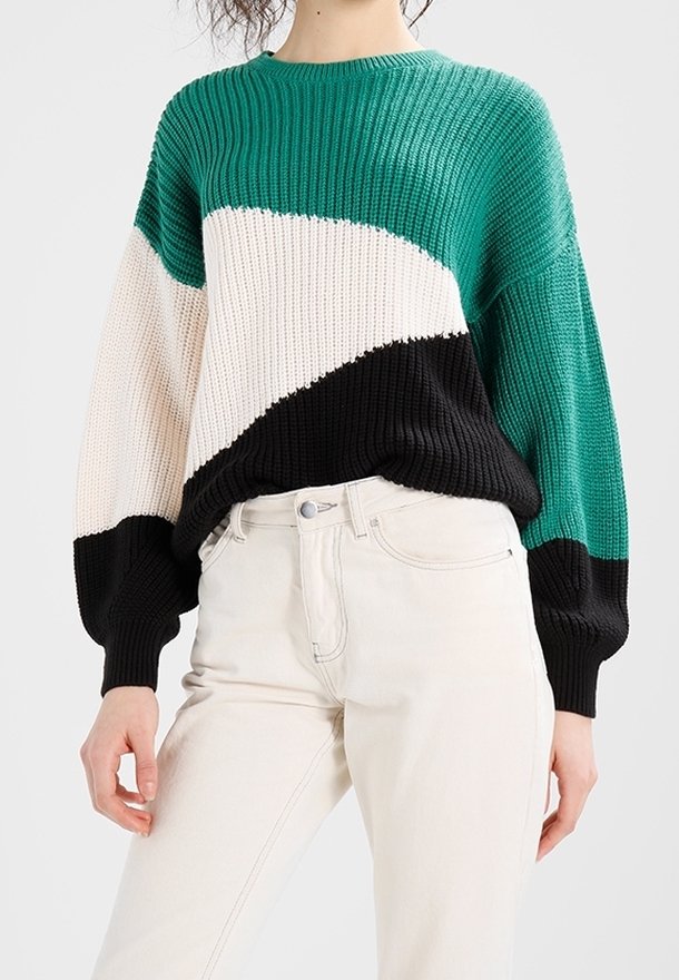 Pull avec des blocs de couleur vert, blanc et noir porté avec un pantalon taille haute blanc cassé. Le haut du corps du mannequin est visible, les bras détendus.