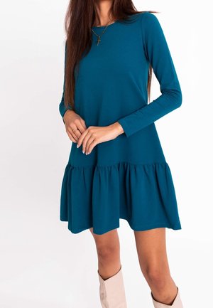 Kobieta w turkusowej sukience z długim rękawem do kolan z falbaną na dole, beżowymi kozakami za kolano i złotym naszyjnikiem z krzyżykiem, stojąca z złączonymi dłońmi.