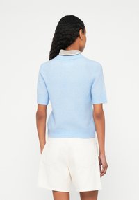 Gaiši zils īsroku džemperis ar ribotu jostasvietu un svītrainu apkakli, kopā ar krēmkrāsas šortiem. Redzama maiga dzijas tekstūra.