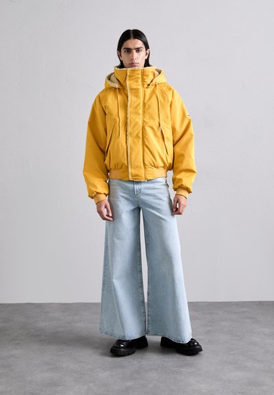 Yellow padded jacket with a high collar, paired with wide-leg light blue jeans. Black chunky shoes complete the outfit. Neutral background.
