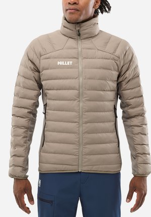 Homme portant une veste matelassée beige avec fermeture éclair et logo « Millet », associée à un pantalon bleu marine avec poche zippée.