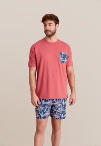 Koraalkleurig shirt met korte mouwen en een geprinte zak; gecombineerd met blauwe shorts met een tropisch bladerenpatroon in rood en wit.