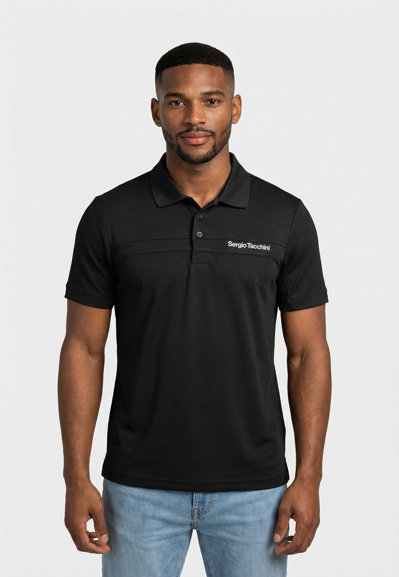 Homme debout, face à l'avant, portant un polo noir Sergio Tacchini et un jean bleu clair, sur un fond uni.