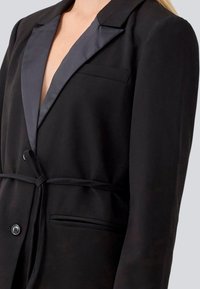 Blazer nero con rever in raso, chiusura con un bottone e cintura in vita. Presenta una tasca sul petto e una texture del tessuto liscia.