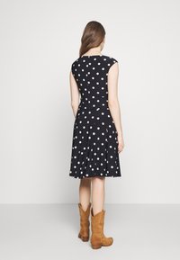 Kvinna som står med ryggen mot kameran, iklädd en ärmlös svart klänning med vita prickar och bruna mocka stövlar som når till halva vadbenet.