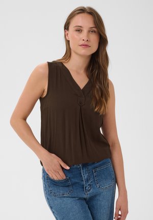 Femme aux longs cheveux bruns portant un chemisier sans manches marron foncé et un jean bleu, debout, une main dans la poche.