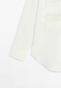 Bílá košile s dlouhým rukávem, která má na přední straně strukturovaný nařasený panel, klasický límec a manžety na knoflíky. Měkký bavlněný materiál.