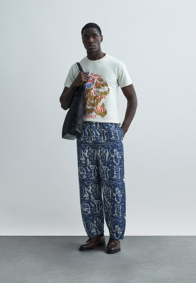 Μπλούζα κρεμ με πολύχρωμο γραφικό λιονταριού; μπλε παντελόνι με φαρδιά πόδια και μοτίβο; καφέ δερμάτινα παπούτσια; μαύρη τσάντα tote. Ορατές λεπτομέρειες υφής υφάσματος.