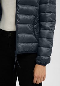 Marineblaue Puffjacke mit glänzender Oberfläche, gesteppten Einsätzen und elastischen Bündchen. Mit Reißverschluss und Seitentasche. Über einem hellen Oberteil getragen.