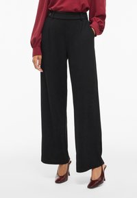 Schwarze weit geschnittene Hosen mit glatter Textur, hoher Taille, vorderen Falten und seitlichen Taschen. Kombiniert mit einer burgunderfarbenen Bluse und braunen Absatzschuhen.