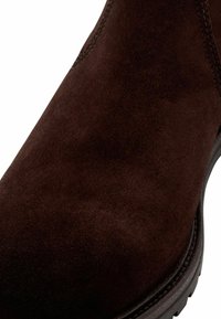 Gros plan sur une botte de cheville en cuir suédé marron foncé avec des coutures visibles et une semelle texturée sur un fond blanc.