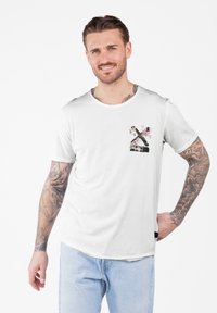 Hombre con brazos tatuados que lleva una camiseta blanca con una X negra y un diseño floral rosa, jeans de color azul claro, sonriendo con una mano en la cadera.